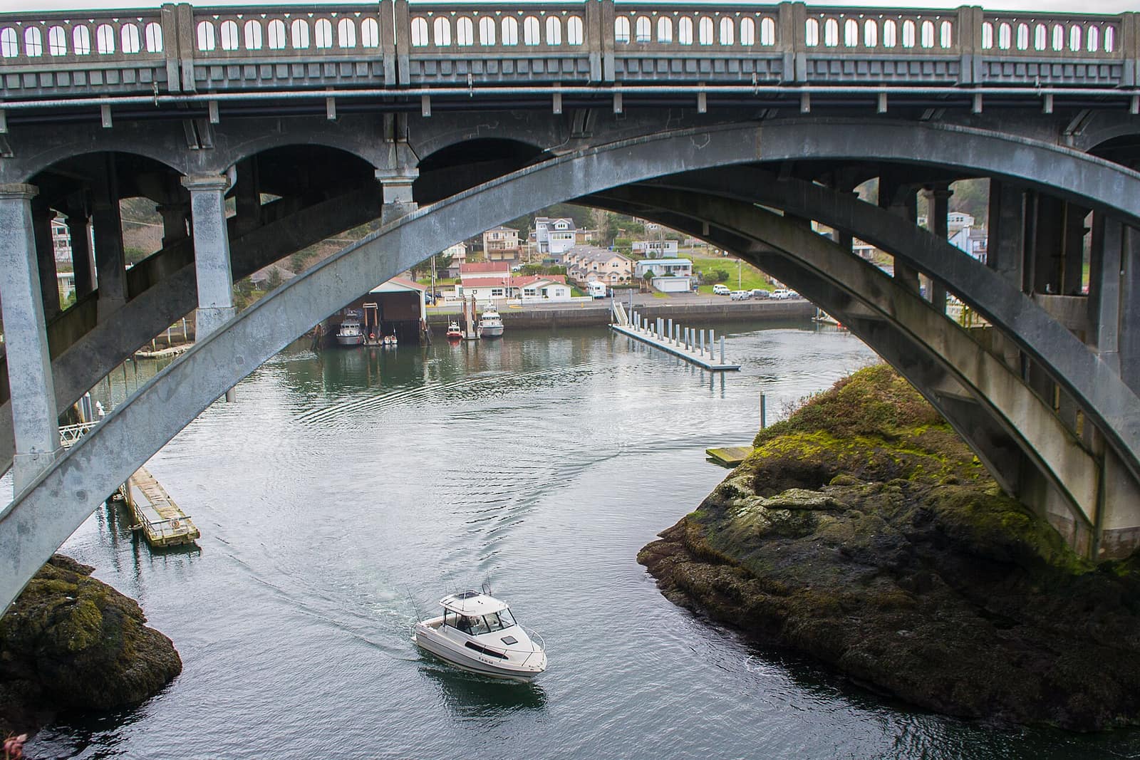 A small white fishing boat passing under the gray concrete arches of the Depoe Bay Bridge, with moorage and houses beyond