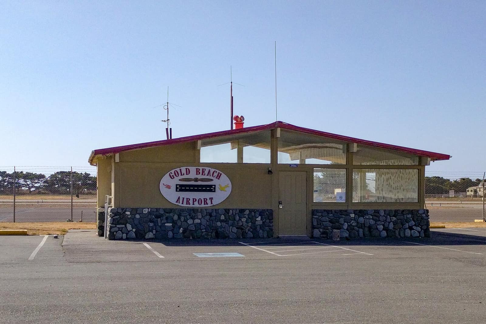 The small office building of the Gold Beach Airport with a stone base and a sign, beside a paved parking lot and the runway