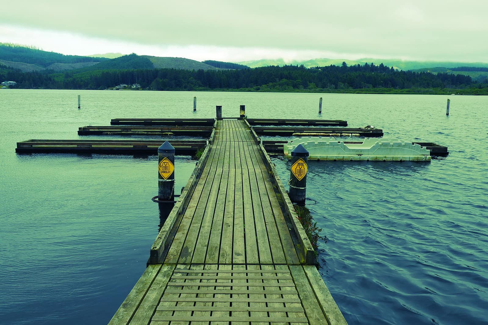 A wooden boat dock extending into Devils Lake in Lincoln City, with forested hills across the water