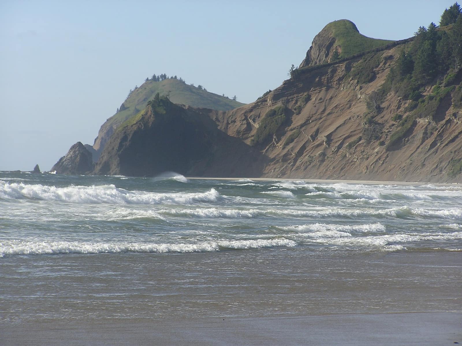 Surf rolling onto the sand at Road&rsquo;s End State Park in Lincoln City, with forested headlands behind