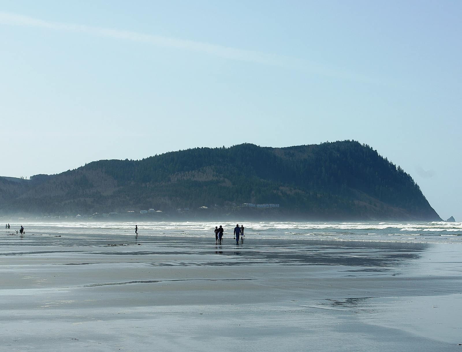 Walkers on a wide, wet beach at Seaside with surf in the distance and the forested bulk of Tillamook Head across the water