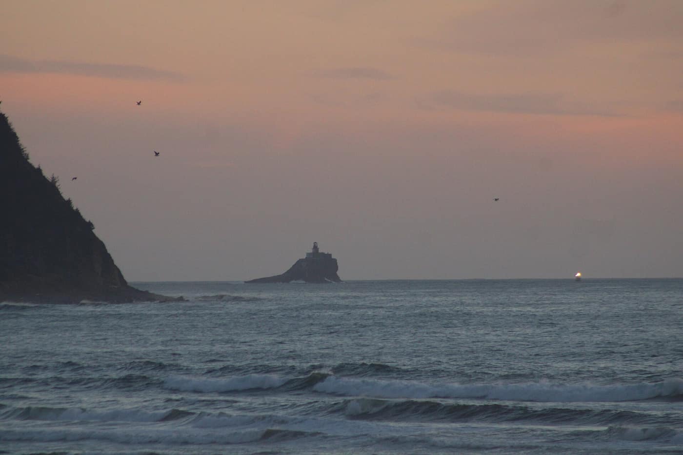 Seaside, Oregon weather at dusk: waves rolling in below Tillamook Head, with Tillamook Rock and its lighthouse beyond